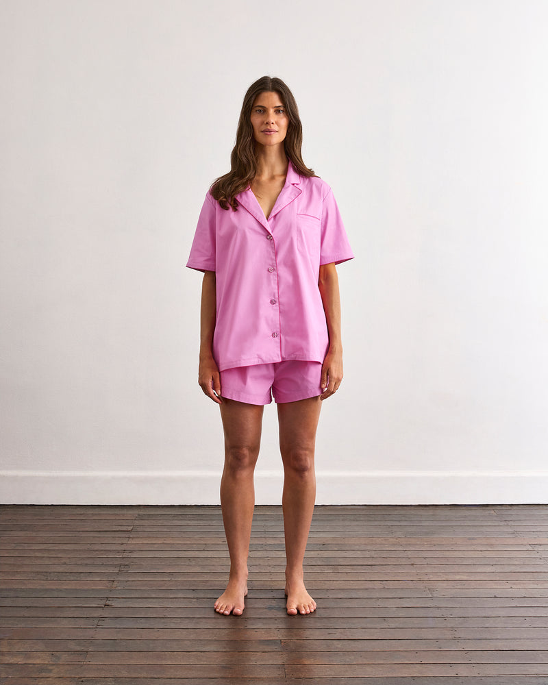 A woman stands barefoot on a wooden floor against a white wall, wearing the Bed Threads Bubblegum & Berry Piped Short Sleep Set—featuring matching pink cotton piped shorts and a short sleeve shirt.