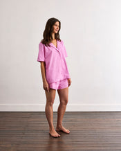 A woman stands barefoot on a wooden floor against a white wall, wearing Bed Threads' Bubblegum & Berry Piped Shorts sleepwear. She faces slightly to the side and smiles gently, showcasing the short-sleeved, loose cotton set. - view