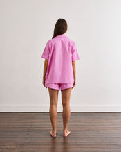 A person with long brown hair stands barefoot on a wooden floor, facing away from the camera, wearing the Bed Threads Bubblegum & Berry Piped Short Sleep Set against a plain white wall. - view