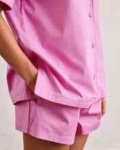 A person models the Bed Threads Bubblegum & Berry Piped Shorts—a loose-fitting, short-sleeved pink button-up shirt and matching piped shorts—standing with one hand in their pocket against a plain background. - view