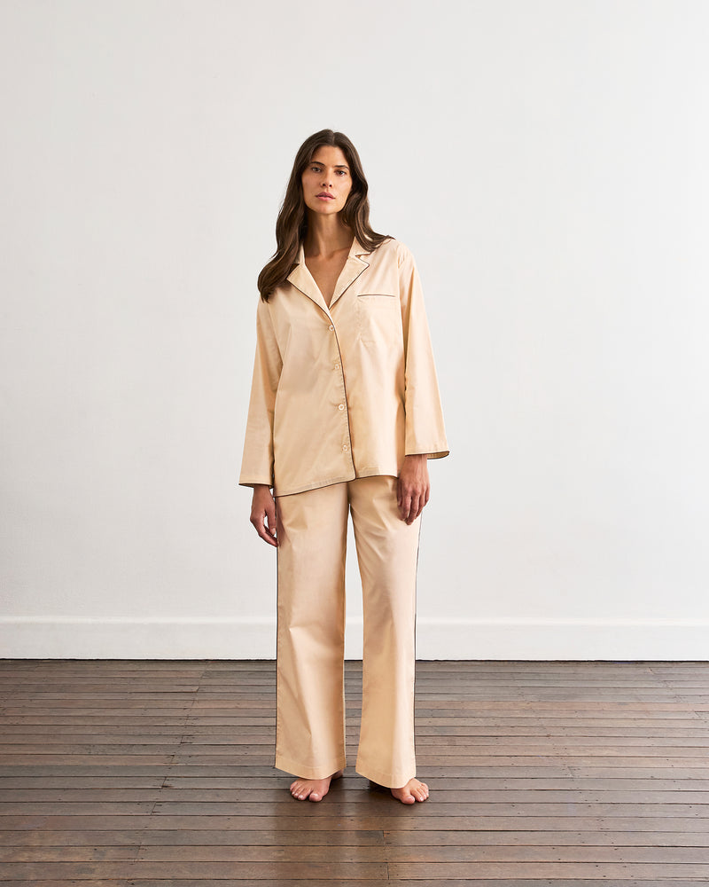 A woman with long brown hair stands barefoot on a wooden floor, wearing the Bed Threads Vanilla & Espresso Piped Long Sleeve Shirt, paired with wide-leg pants, against a plain white wall.