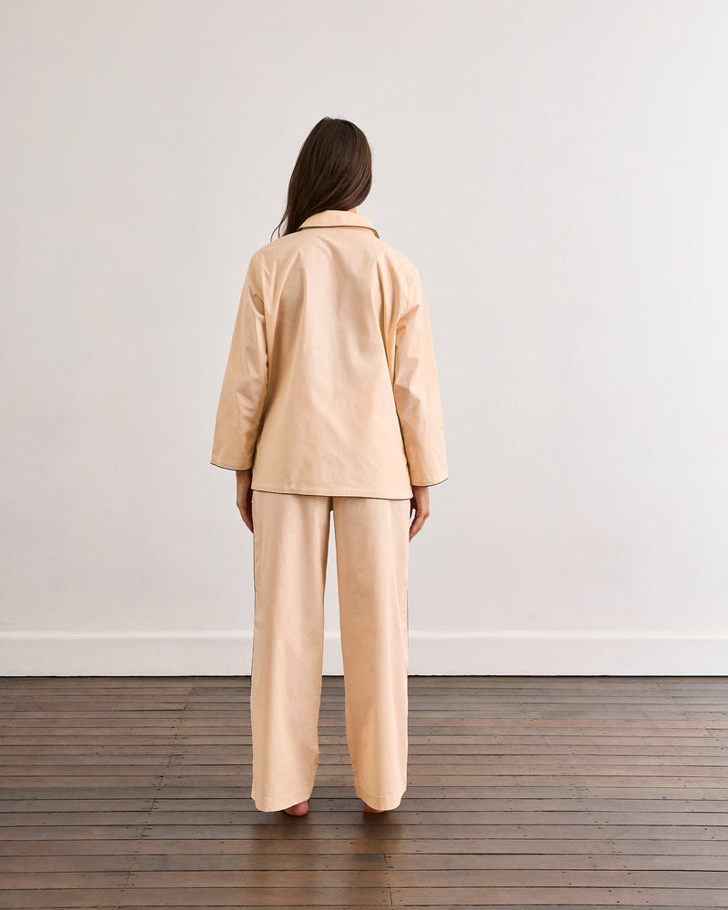 A person with long hair stands barefoot on a wooden floor, facing a plain white wall, wearing the Bed Threads Vanilla & Espresso Piped Long Sleeve Shirt.