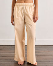A person stands barefoot on a wooden floor, wearing loose, light beige pajama pants from the Vanilla & Espresso Piped Long Sleep Set by Bed Threads. The photo is cropped to show only the lower half of their body. - view
