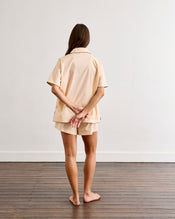 A woman with long, loose hair stands barefoot on a wooden floor facing a white wall, hands behind her back. She wears the Bed Threads Vanilla & Espresso Piped Short Sleeve Shirt and matching shorts, creating a relaxed look. - view