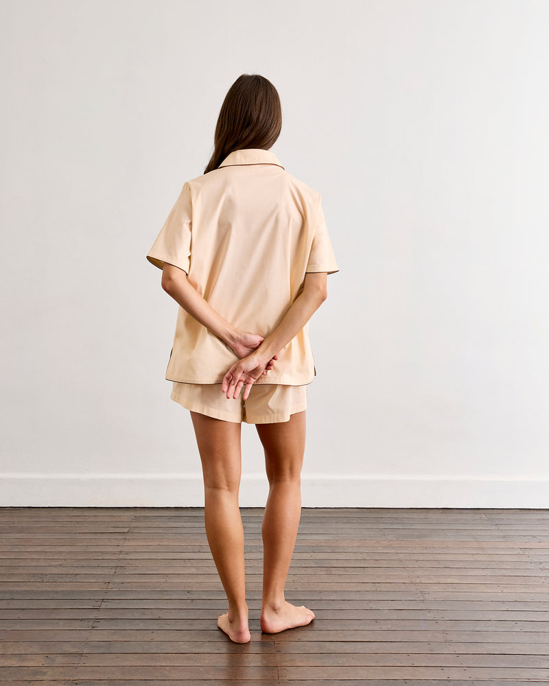 A woman stands barefoot on a wooden floor facing a white wall, hands behind her back. She wears the Vanilla & Espresso Piped Short Sleep Set by Bed Threads—a lightweight cotton set with a short sleeve shirt and matching piped shorts in beige.