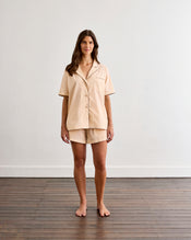 A woman stands barefoot on a wooden floor in front of a white wall, wearing the Bed Threads Vanilla & Espresso Piped Short Sleep Set—a lightweight cotton short-sleeve shirt and piped shorts. - view