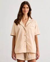 A woman with long brown hair wears the Bed Threads Vanilla & Espresso Piped Short Sleeve Shirt, a lightweight cotton set with contrast piping, standing against a plain light background and smiling slightly. - view