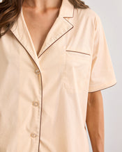 A person wears the Bed Threads Vanilla & Espresso Piped Short Sleeve Shirt, a lightweight cotton top in light beige with dark brown piping on the collar, pocket, and sleeves. The shirt has a chest pocket and an open collar. - view