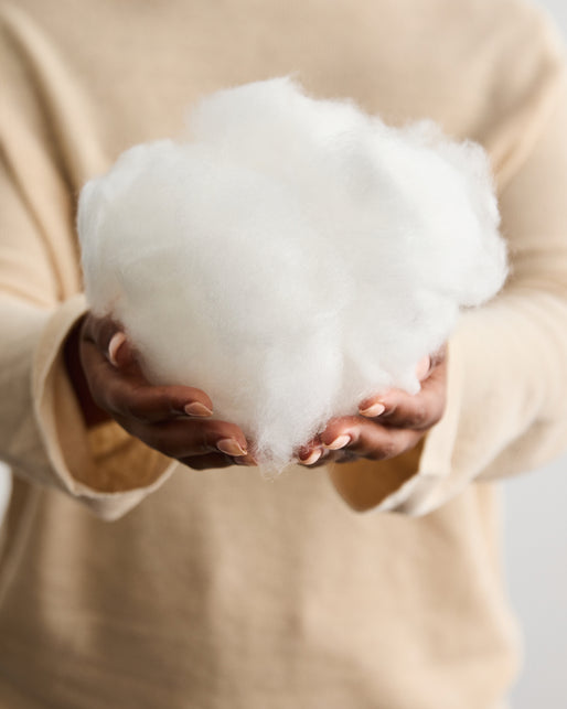 A person wearing a beige sweater holds a fluffy white ball of cotton or fiberfill in their hands, presenting it toward the camera against a neutral background.