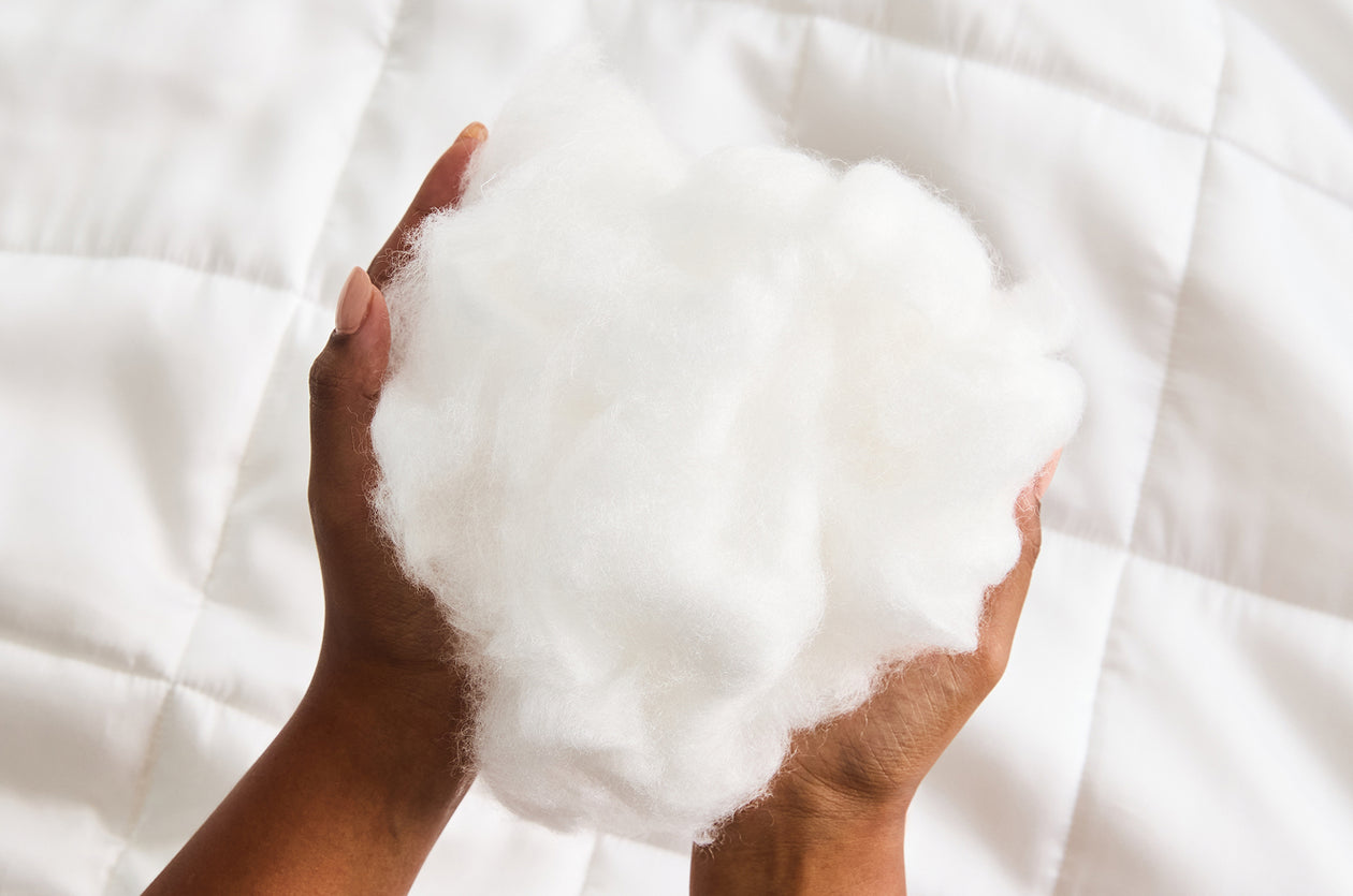 Two hands hold a fluffy, white bundle of synthetic fiber filling over a white, quilted fabric background.