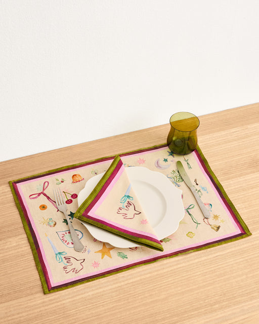 A table setting with a white plate, Bed Threads La Fête 100% French Flax Linen Napkins (Set of Four), a whimsical placemat, cutlery, and an olive-green glass on light wood—a festive decor choice.