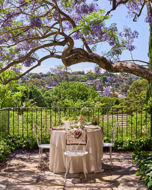 A round table with festive decor and Bed Threads' La Fête 100% French Flax Linen Napkins (Set of Four) sits under a blooming jacaranda on a sunny terrace, overlooking a green garden and distant hills.