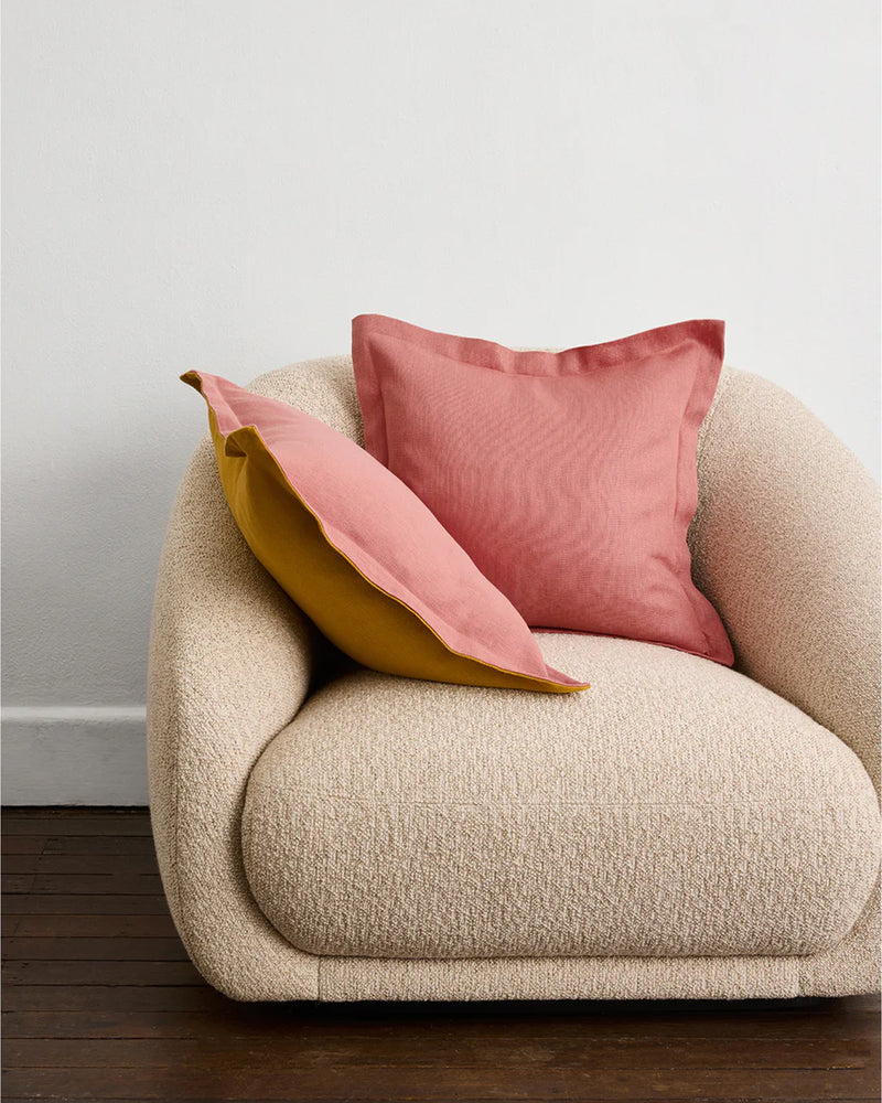 A plush cream armchair with two square cushion covers—one Pink Clay & Turmeric 100% French Flax Linen Cushion Cover by Bed Threads—stands on wooden flooring against a plain white wall.