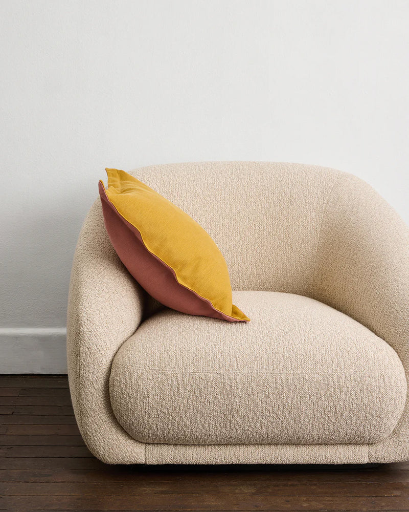 A cream-colored, rounded armchair featuring a Bed Threads Pink Clay & Turmeric 100% French Flax Linen Cushion Cover rests on a dark wooden floor against a plain white wall.
