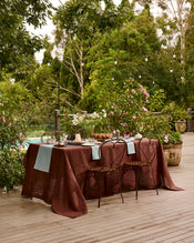 A Lagoon 100% French Flax Linen Table Runner by Bed Threads adorns a rustic outdoor table on a wooden deck, set for dining amid lush greenery and flowers, with tableware and string lights creating an inviting atmosphere. - view