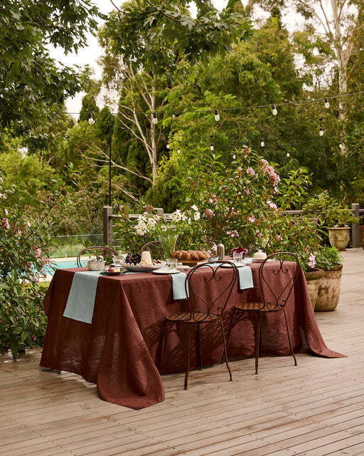 A Lagoon 100% French Flax Linen Table Runner by Bed Threads adorns a rustic outdoor table on a wooden deck, set for dining amid lush greenery and flowers, with tableware and string lights creating an inviting atmosphere.