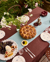 An outdoor meal is set with a brown tablecloth and a Lagoon 100% French Flax Linen Table Runner by Bed Threads, featuring bread, figs, grapes, cheese, placemats, white flowers in a vase, plates, silverware, and glasses. - view