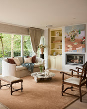 A cozy living room featuring a Bed Threads Lagoon & Crème 100% French Flax Linen Muslin Throw, a white linen sofa with pillows, marble coffee table with flowers, armchair, fireplace, art, decor shelves, and large windows overlooking green trees. - view