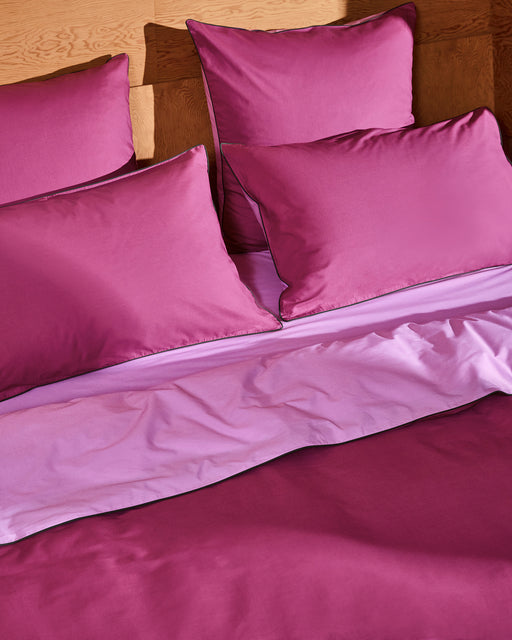 A neatly made bed with magenta and light purple bedding features four large pillows against a wooden headboard, dressed in Bed Threads Berry, Bubblegum & Moss Piped 100% Organic Cotton European Pillowcases for a soft, luxurious feel.