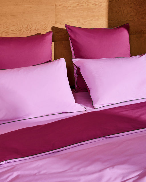A neatly made bed with Berry, Bubblegum & Moss Piped 100% Organic Cotton European Pillowcases (Set of Two) by Bed Threads sits against a wooden headboard, sunlight partially illuminating the light pink and magenta bedding.