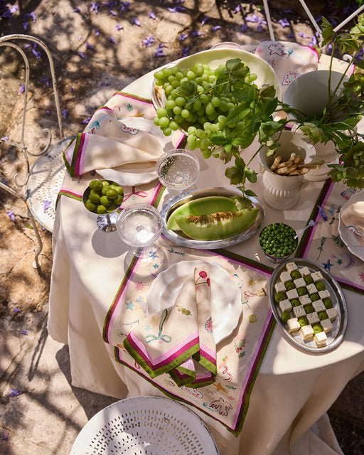 An outdoor table is set with a cream cloth, Bed Threads La Fête 100% French Flax Linen Napkins (Set of Four), grapes, olives, melon, petit fours, and a leafy centerpiece. Sunlight and purple petals enhance the festive decor.