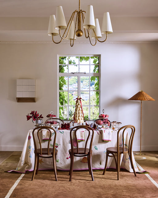 A dining table set for celebration is adorned with the La Fête 100% French Flax Linen Tablecloth by Bed Threads, topped with desserts and a croquembouche, beneath a white and gold chandelier. Wooden chairs and a leafy view complete the scene.