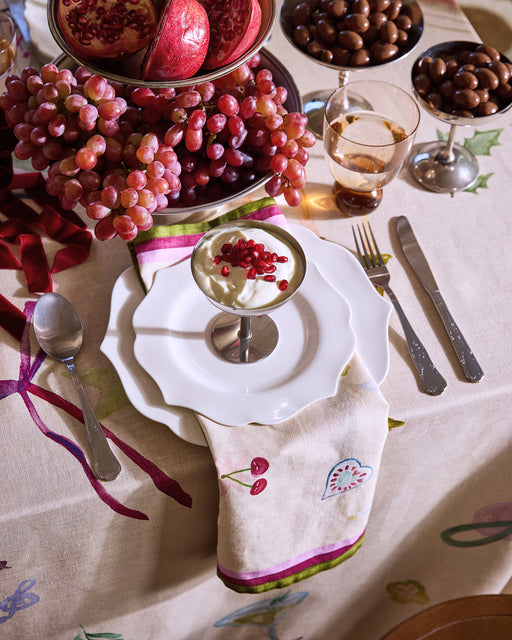 A festive table features Bed Threads’ La Fête 100% French Flax Linen Napkins (Set of Four), a white plate, silver dessert dish with pomegranate seeds, water glass, cutlery, red grapes, whole pomegranate, and chocolate-covered treats.