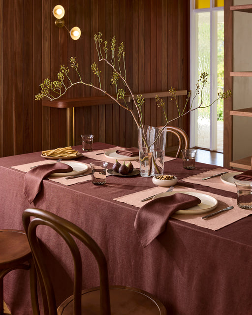A dining table set with a burgundy cloth, Bed Threads’ Hazelnut 100% French Flax Linen Placemats, matching napkins, white plates, glasses, and green branches as a centerpiece. Wood-paneled walls and wooden chairs frame the arrangement.