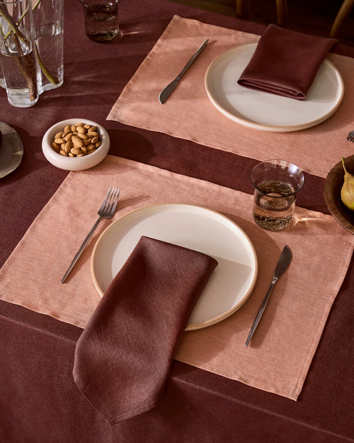 A table set for two features Bed Threads Hazelnut 100% French Flax Linen Placemats, white plates, burgundy napkins, silver cutlery, and water glasses. A bowl of nuts and a vase with branches add charm atop a dark burgundy tablecloth.