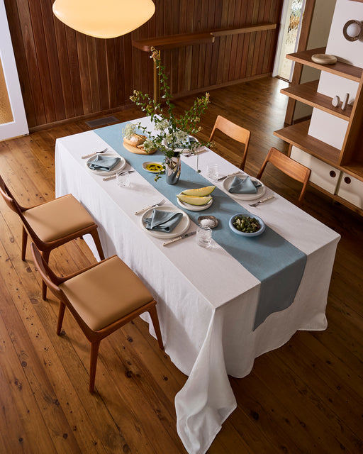 A dining table with a white tablecloth and the Bed Threads Lagoon 100% French Flax Linen Table Runner is set with plates, glasses, napkins, cutlery, a vase of flowers, and fruit bowls. Four wooden chairs are arranged on the wooden floor.