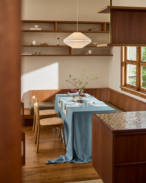 A modern dining room features natural wood furniture, built-in benches, open shelves, wooden floors, large windows, and a Bed Threads Lagoon 100% French Flax Linen Tablecloth draped over the table set for a meal.