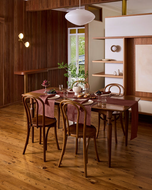 A cozy dining room with a wooden table set for four, featuring brown chairs and Bed Threads Cacao 100% French Flax Linen Placemats (Set of Four), elegant tableware, lush plants, wooden floors, and modern lighting for a warm, inviting feel.