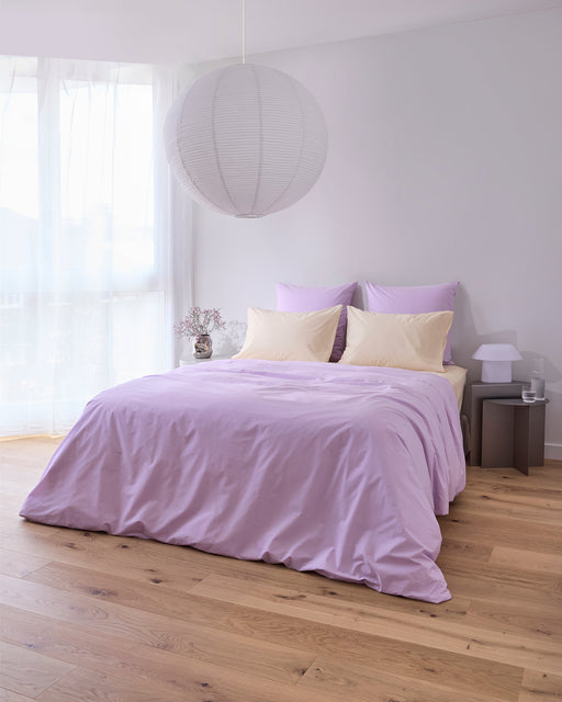 A neatly made bed with the Bed Threads Violet 100% Organic Cotton Duvet Cover and cream pillows sits in a bright, minimalist bedroom with light wood floors, a large paper lantern ceiling light, and soft natural light through sheer curtains.