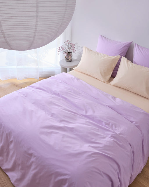 A neatly made bed with Bed Threads’ Violet & Vanilla 100% Organic Cotton Bedding Bundle and cream pillows sits by a round nightstand with flowers, while a large paper lantern near sheer white curtains fills the room with soft light.