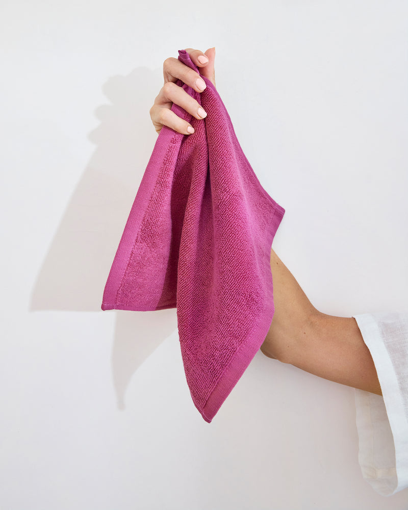 A hand holds a neatly folded, bright pink Berry 100% Cotton Terry Face Washer from Bed Threads (set of two) against a plain white background.
