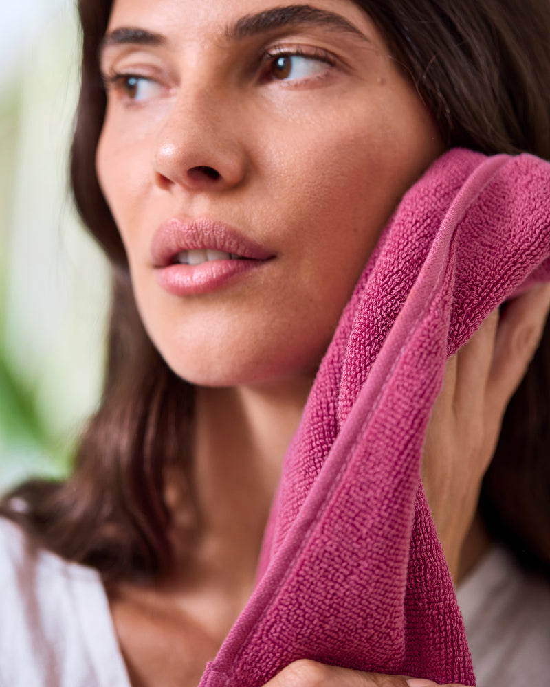A woman with long brown hair gently presses a Berry 100% Cotton Terry Face Washer by Bed Threads to her cheek. Calm and wearing a white top, she looks off to the side, showcasing this soft, absorbent set of two face washers.