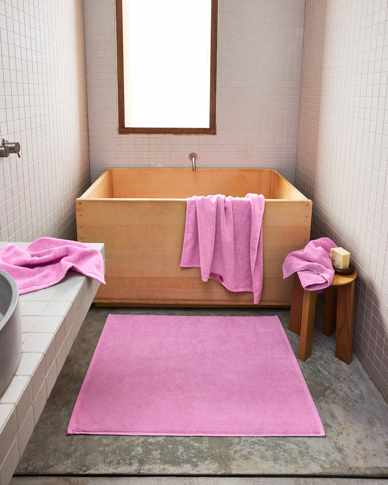 A minimalist bathroom features a light wooden tub, a pink towel on its edge, and the Bed Threads Bubblegum 100% Cotton Terry Bath Mat on the floor, with another pink towel and soap resting on a small wooden stool.