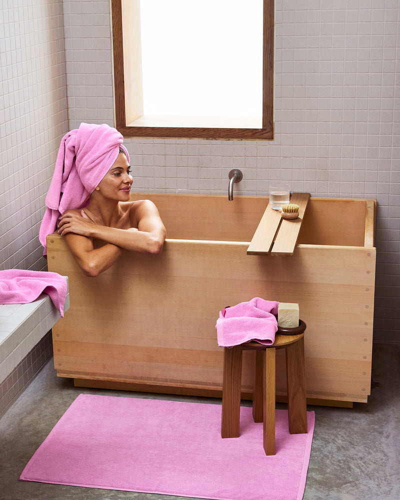 A person relaxes in a wooden tub with a pink Bubblegum 100% Cotton Terry Bath Sheet by Bed Threads wrapped around their head, plush quick-drying towels and a bath mat nearby, while a stool holds soap and water.