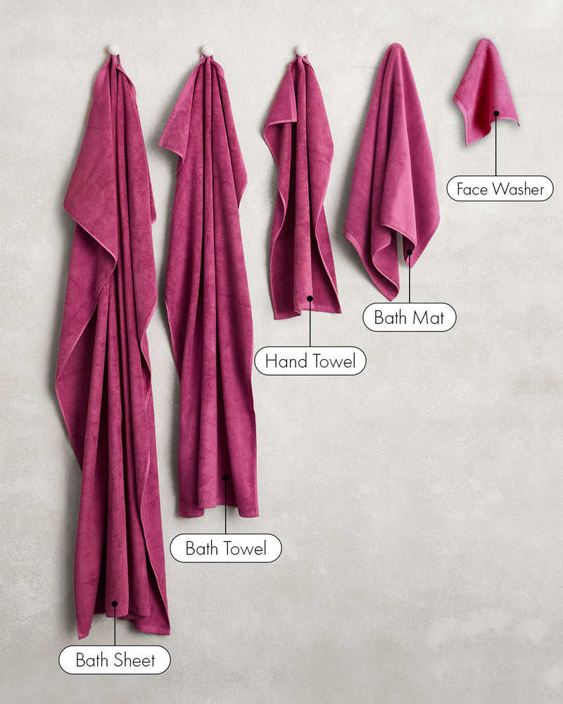 Five magenta towels from Bed Threads, including a Bath Sheet, Bath Towel, Hand Towel, Bath Mat, and a set of Berry 100% Cotton Terry Face Washers (two included), hang on hooks against a light wall.