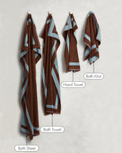 Two Cacao & Lagoon Two-Toned 100% Cotton Terry Hand Towels by Bed Threads hang on hooks against a light wall—a stylish set that adds elegance and function to any bathroom. - view