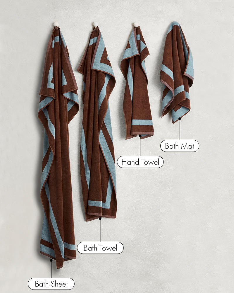 Two Cacao & Lagoon Two-Toned 100% Cotton Terry Hand Towels by Bed Threads hang on hooks against a light wall—a stylish set that adds elegance and function to any bathroom.