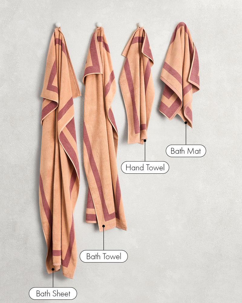 A Bed Threads Terracotta & Pink Clay Two-Toned 100% Cotton Terry Hand Towel, with plush 650GSM softness and quick-drying performance, hangs on a light wall—displaying its elegant peach and maroon design.