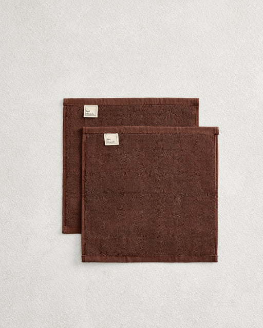 The Bed Threads Espresso 100% Cotton Terry Face Washers (Set of Two) feature dark brown rectangles with stitched edges and fabric labels, layered on a light surface. Made from absorbent terry, they're gentle on skin.