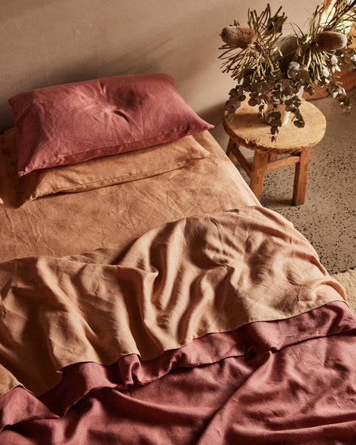 A cozy bed dressed in the Bed Threads Pink Clay & Terracotta 100% French Flax Linen Bedding Bundle sits beside a rustic wooden stool topped with a dried floral arrangement as sunlight casts warm shadows over the softly rumpled sheets.