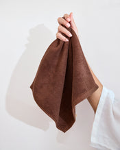 A hand with light pink nails holds up a Bed Threads Espresso 100% Cotton Terry Face Washer (Set of Two), gentle and absorbent, against a plain light background. - view