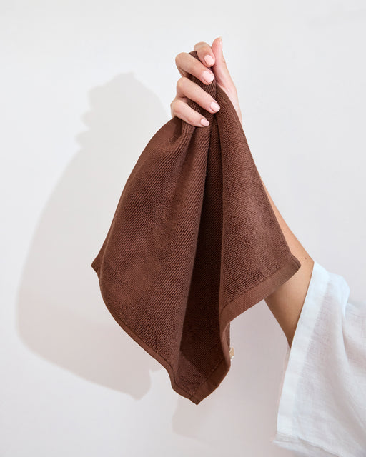 A hand with light pink nails holds up a Bed Threads Espresso 100% Cotton Terry Face Washer (Set of Two), gentle and absorbent, against a plain light background.