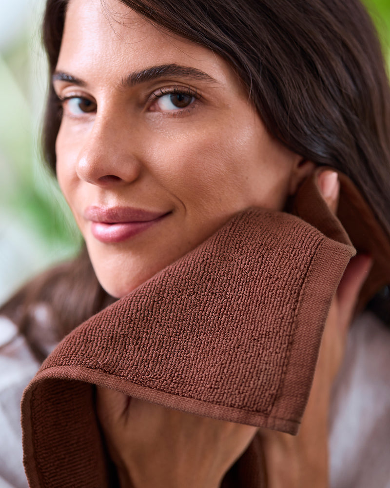 A woman smiles softly as she dries her face with a Bed Threads Espresso 100% Cotton Terry Face Washer (Set of Two), its brown, absorbent fabric complementing the calm, fresh atmosphere and providing gentle care for her skin.