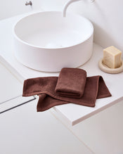 A folded Espresso 100% Cotton Terry Face Washer by Bed Threads sits on a white bathroom countertop beside a round sink and soap dish. The absorbent, gentle terry adds comfort and style to the modern, minimal setting. Set of two. - view