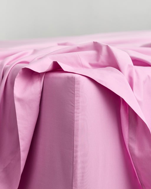 A neatly made bed features Bubblegum Organic Cotton King Pillowcases (Set of Two) by Bed Threads, set against a plain white wall on a dark wooden floor. The smooth, slightly wrinkled bedding adds charm and cool comfort.