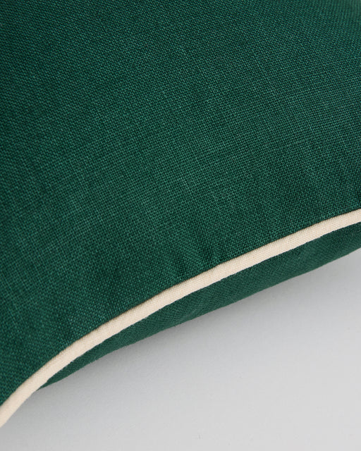A close-up of the Bed Threads Forest & Crème 100% French Flax Linen Piped Cushion Cover, in green with white piping, sits on a light grey surface.